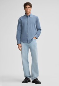 Lichtblauwe overhemden gemaakt van zachte stof, met lange mouwen, een kraag en zichtbare knopen; gedragen met lichtblauwe jeans en zwarte schoenen.