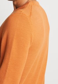 Pull en maille texturée orange avec un design à côtes, présentant un col rond et des détails de couture visibles aux épaules.