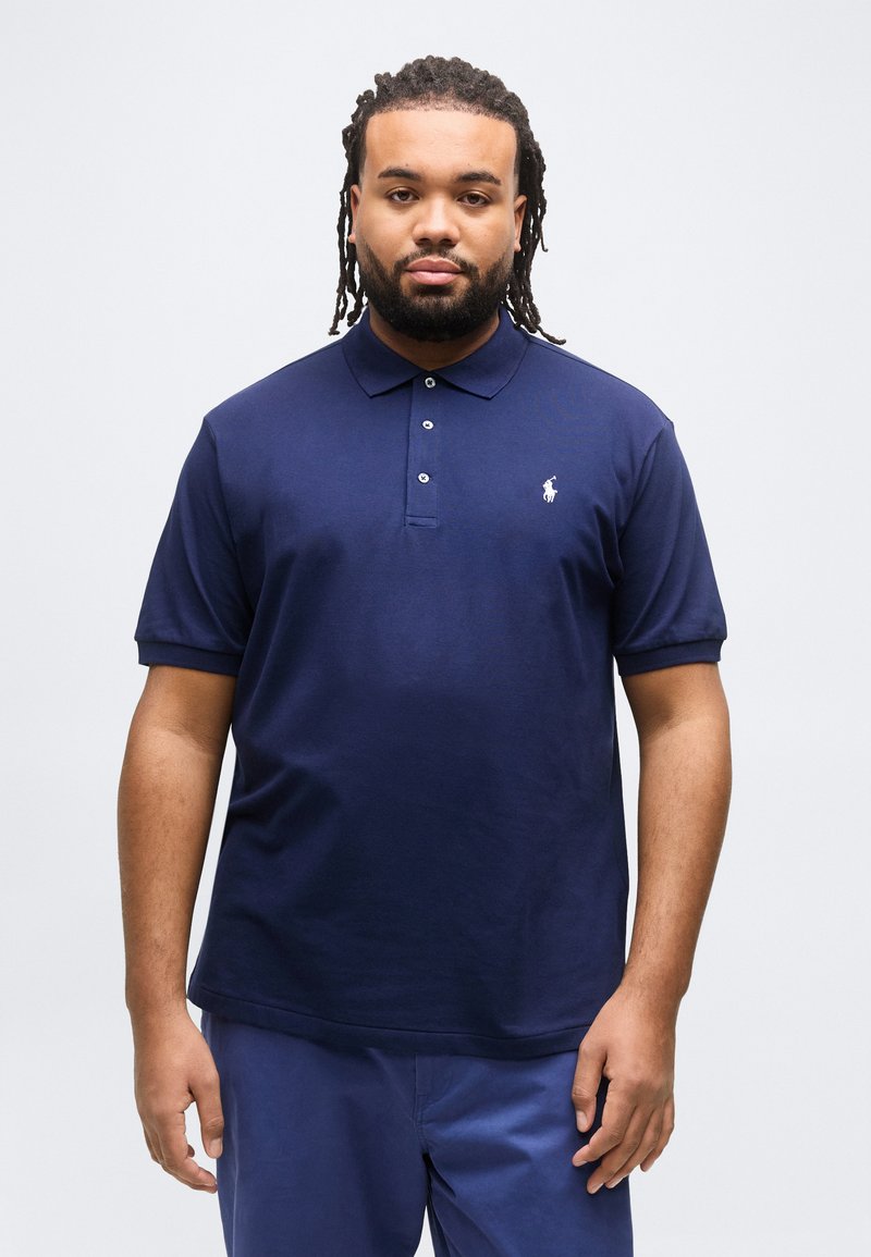 Άνδρας με πλεξούδες στα μαλλιά και γένια, φορώντας μπλε σκουρόχρωμο πόλο πουκάμισο και αντίστοιχο παντελόνι, στέκεται μπροστά σε μια απλή φωτεινή επιφάνεια.