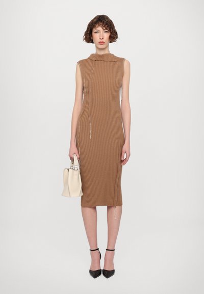 Stickad, ärmlös klänning i beige, med en ribbad textur, asymmetrisk krage och sid sömmar. Modellen håller en liten vit handväska.