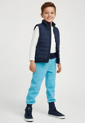 Uśmiechnięty chłopiec stoi, ubrany w granatową pikowaną kamizelkę, białą koszulę z długim rękawem, jasnoniebieskie spodnie dresowe, granatowe skarpetki i granatowe trampki za kostkę.