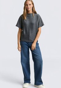 Grå metallisk kortärmad topp med lös passform, kombinerad med blå jeans med vida ben och vita sneakers. Neutral bakgrund.