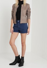 Veste bomber en cuir marron, col roulé noir, shorts en denim et bottines noires. Texture lisse avec un design ajusté et un style décontracté.