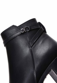 Gros plan d'une botte cheville en cuir noir avec une sangle décorative et une boucle métallique sur le côté supérieur.