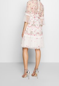 Femme portant une robe blanche transparente mi-longue avec une broderie florale rose et des sandales argentées à talons hauts avec bride à la cheville, vue de dos.