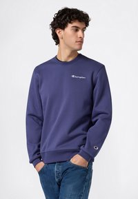 Joven con cabello rizado vistiendo una sudadera azul de Champion y jeans, con las manos en los bolsillos, mirando hacia su derecha contra un fondo simple.