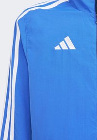 Blå vindjacka med vita ränder längs ärmarna och en vit logotyp på bröstet. Tillverkad av lätt, texturerad tyg.