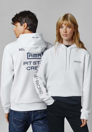 Hombre y mujer vistiendo sudaderas blancas de Red Bull Racing, la mujer mirando hacia adelante, el hombre de espaldas con el texto "Pit Stop Crew".