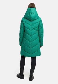 Zielona kurtka puchowa z karbowanym wzorem w zygzak i dużym kapturem, noszona na czarnych spodniach i masywnych czarnych botkach.