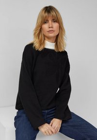 Maglione nero a maniche lunghe con motivo floreale testurizzato, collo rotondo, indossato sopra un dolcevita bianco, abbinato a jeans blu.