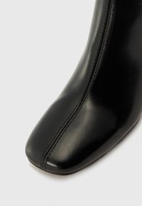 Bottine en cuir noir à bout carré, avec un détail de couture centrale et une texture lisse et brillante.
