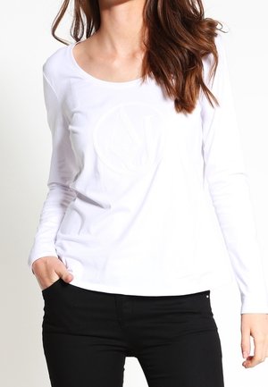 Žena oblečená v bílé halence s dlouhým rukávem a jemným kruhovým logem, černými kalhotami a s jednou rukou v kapse, stojící před jednobarevným pozadím.