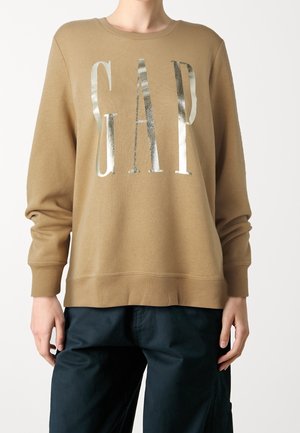 Osoba ubrana w beżową bluzę z długim rękawem z błyszczącymi srebrnymi literami "GAP" z przodu oraz ciemne spodnie, stojąca na jednolitym tle.