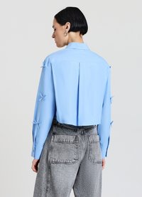 Blouse cropped bleu clair à manches longues, avec des nœuds en tissu aux poignets et une pince dans le dos. Associée à un denim gris taille haute.