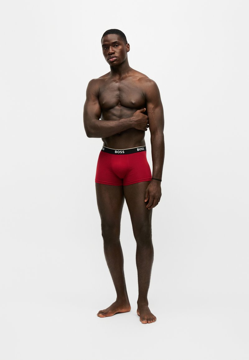 Homme musclé debout pieds nus portant un boxer rouge BOSS, avec un bras croisé sur la poitrine sur un fond blanc uni.