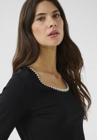 Juoda, raukšlėta ilgomis rankovėmis palaidinė su baltu banguotu apvadu ties kaklu. Audinys atrodo lygus, su prigludusia siluetu.