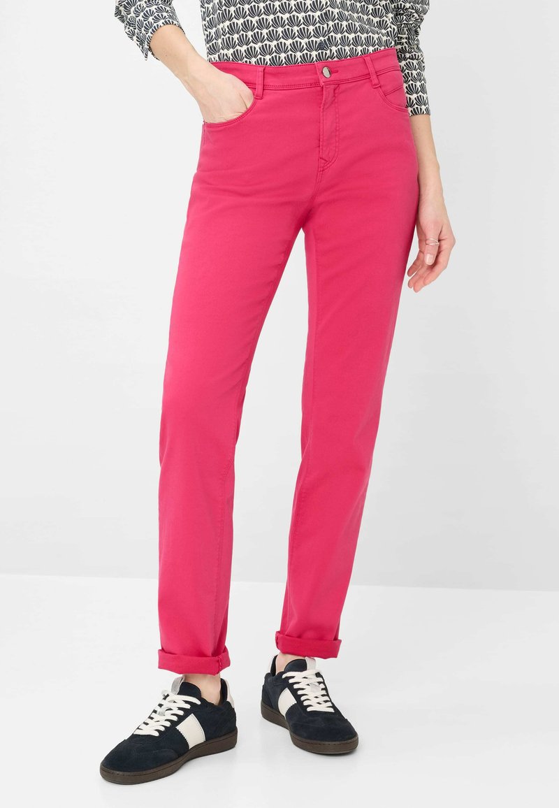 Person trägt knallig pinke, eng anliegende Hosen mit aufgerollten Säumen, schwarz-weiße Turnschuhe und ein gemustertes langärmliges Oberteil, steht vor weißem Hintergrund.