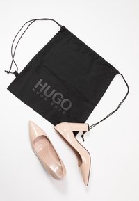 Talons hauts en cuir verni beige avec des pointes, accompagnés d'un sac à cordon noir arborant un logo gris. Le talon présente un design unique.