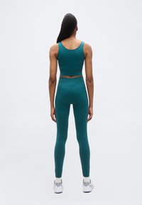 Γυναίκα που στέκεται πλάτη φοράει σετ γυμναστικής σε τιρκουάζ ριμπ ύφασμα, με crop top και ψηλόμεσα κολάν, λευκά αθλητικά παπούτσια και κάλτσες σε απλό φόντο.