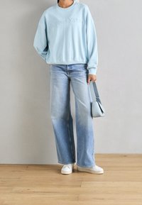 Light blue sweatshirt with embroidered logo, paired with light wash wide-leg jeans and white sneakers. Carrying a small light blue bag.
