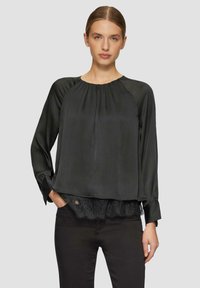 Zwarte blouse met lange mouwen, geplooide textuur, ronde halslijn en kantafwerking aan de zoom, gecombineerd met een donkere broek.