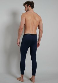 Shirtless man standing barefoot, facing away, wearing fitted dark blue long underwear pants with a black waistband.