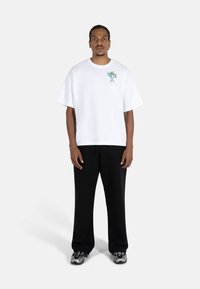 Valge puuvillane T-särk lõdva lõikega, lühikeste varrukatega ja vasakul rinnal värvikas graafiline kujundus. Kombineeritud mustade laia säärega pükstega.