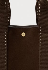 Σκούρο καφέ υφασμάτινο σακίδιο με φαρδιά ραμμένα λουριά και χρυσό κουμπί φτάρνι στο κεντρικό επάνω μέρος.