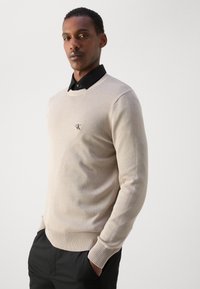 Beige pulóver kerek nyakkivágással, bordás aljúval és mandzsettával, valamint egy kis fekete "CK" logóval a bal mellen; fekete galléros ing fölött viselve.