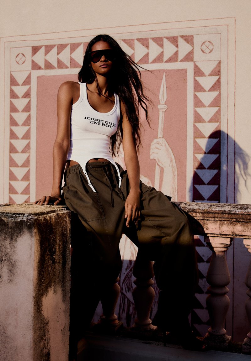 Femme portant des lunettes de soleil, un débardeur blanc avec un texte et un pantalon cargo foncé, assise sur une rampe en pierre contre une fresque murale décorative au soleil.