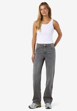 Tânără purtând un top alb fără mâneci cu dungi, blugi gri cu croială dreaptă și pantofi sport argintii, stând cu o mână pe spate.