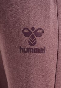 Sittande, mjuk, lila tyg med en mörklila logotyp med ett bihmönster, placerad nära midjan på benet av plagget.
