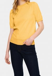 Žlutý pulovr s krátkým rukávem a kulatým výstřihem, s vroubkovanými manžetami a strukturovaným pletem, v kombinaci s tmavými džíny.