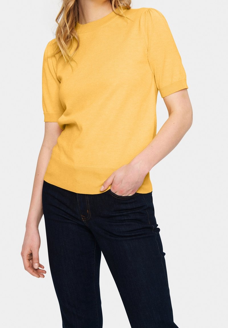 Žlutý pulovr s krátkým rukávem a kulatým výstřihem, s vroubkovanými manžetami a strukturovaným pletem, v kombinaci s tmavými džíny.