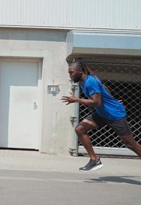 Läufer in einem blauen Kurzarmshirt und schwarzen Shorts, mit schwarzen Sportschuhen, der auf einer asphaltierten Fläche nahe einer strukturierten Wand sprintet.