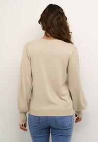 Beige adīta džemperis ar garām, pūkainām piedurknēm un ribotām aprocēm. Ar noapaļotu dekoltē un pieguļošu apakšmalu, kombinēts ar ziliem džinsiem.