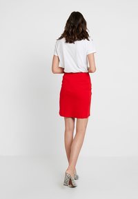 Jupe rouge ajustée avec une texture lisse et une taille, associée à un t-shirt blanc uni et des talons à motifs, vue de l'arrière.