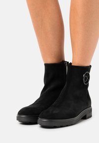 Bottines en daim noir avec une semelle en caoutchouc plate, dotées d'une fermeture éclair latérale et d'un accent logo circulaire. Texture lisse avec un design élégant.