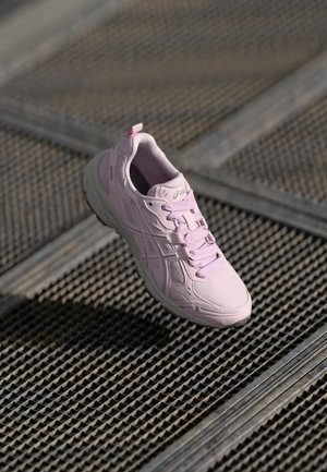 Baskets ASICS rose clair avec lacets flottant au-dessus d'un sol en grille métallique dans une lumière naturelle douce.