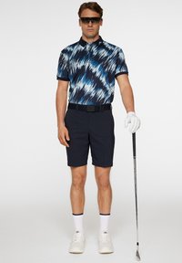 Polohemd mit blauem und navy-farbenem abstraktem Muster; dunkle Shorts; weiße Sneaker; weißer Golfschuh; hält einen Golfschläger; Sonnenbrille.