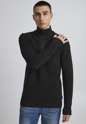 Black ribbed sweater with a high collar and half-zip closure. Features long sleeves with extended cuffs. Worn with blue jeans.
