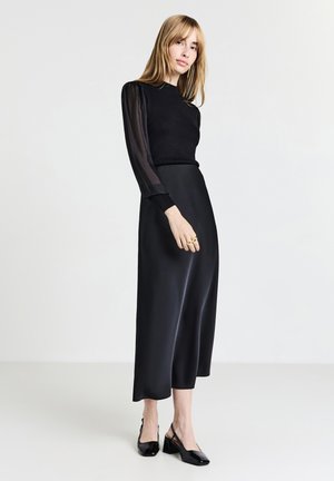 Top nero a maniche lunghe con maniche trasparenti, abbinato a una gonna di satin nera. La modella indossa scarpe slingback nere con finitura lucida.