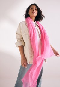 Rosa Schal aus leichtem, strukturiertem Stoff, der elegant um den Hals drapiert ist; getragen zu einer hellen Jacke und Jeans.