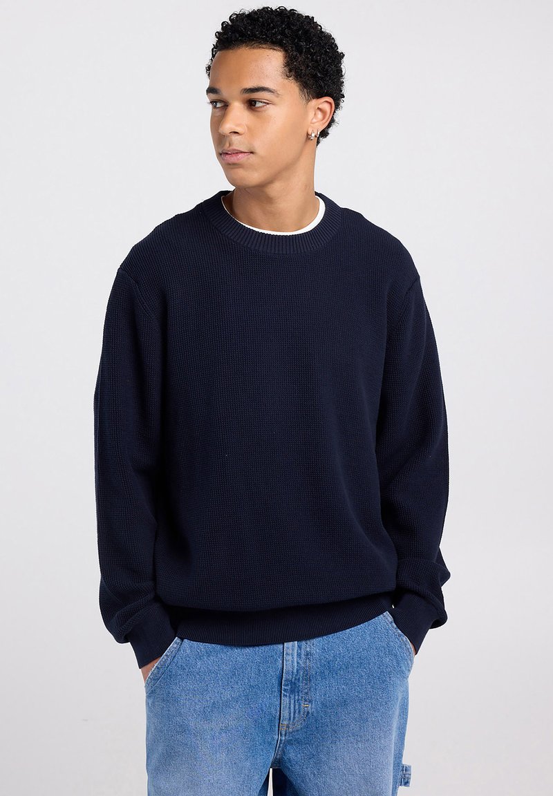 Giovane uomo con capelli ricci che indossa un maglione blu navy e jeans azzurri, guarda verso sinistra con le mani in tasca.