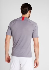 Šedé sportovní tričko s krátkými rukávy, přiléhavým střihem a červeným akcentem na zadním výstřihu. Texturovaná tkanina pro prodyšnost.