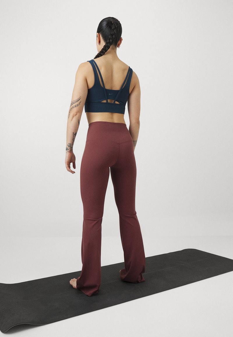 Marineblauw sportbh met een ontwerp van meerdere bandjes aan de achterkant, gecombineerd met bordeauxrode flared legging, staand op een zwarte yogamat.