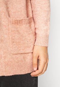 Světle růžový pletený kardigan s žebrovaným lemem a manžetami, s detailem přední kapsy. Textura působí měkce a načechraně.