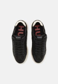 Black athletic shoes with a textured upper, lace closure, logo tag, and contrasting cream accents on the heel and sole.