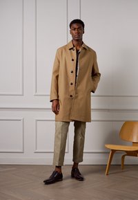 Abrigo trench caqui con botones oscuros, que presenta un corte recto y bolsillos laterales, combinado con pantalones beige claros y zapatos de cuero marrón.