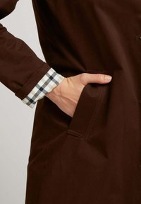 Manteau marron présenté avec une main dans la poche latérale ; la manche comporte un motif à carreaux noir et blanc au niveau du poignet.